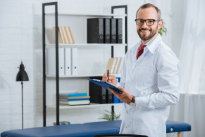 portrait of smiling male chiropractor in white coat and eyeglasses with notepad in hospital