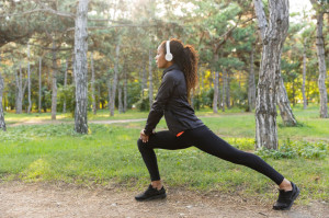 Image of sportswoman 20s wearing black tracksuit working out, and stretching body in green park